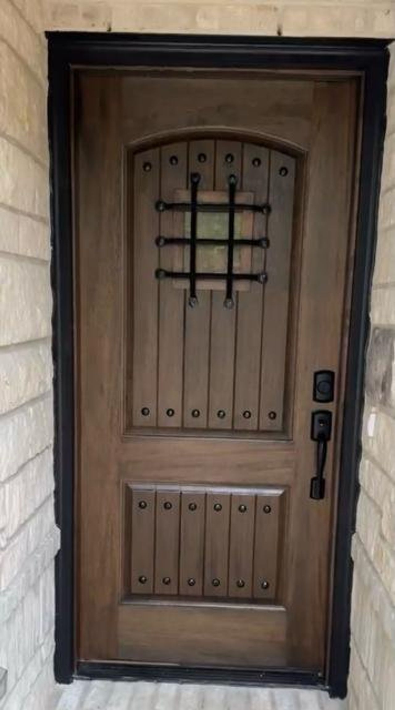Arched wood-finish entry door featuring decorative iron-grille window, black hardware, and studded accents