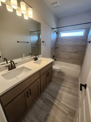 A modern bathroom with wood cabinetry, dual sinks, sleek lighting, and a tiled bathtub.