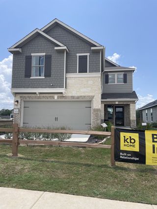 Street view A spacious two-story home in Mission del Lago by KB Home (San Antonio, TX), featuring a stone and gray siding exterior, a two-car garage, and a "Built on Relationships" sign.