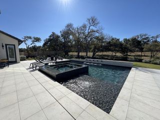 Model Home A modern backyard oasis featuring a dark tiled swimming pool with a built-in spa, lounge chairs, and a fire pit area, surrounded by a landscaped yard and mature trees.
