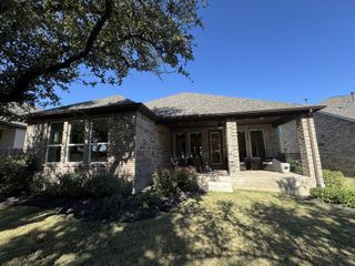 A well-maintained backyard featuring a single-story brick home with a covered patio area. The outdoor space is landscaped with greenery and shaded by mature trees.