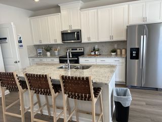 A bright, modern kitchen with white cabinetry, stainless steel appliances, a granite-topped island with seating, and stylish woven bar stools.