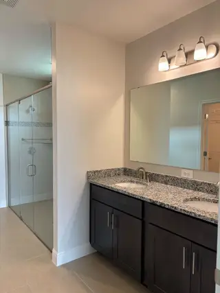 A modern bathroom featuring a granite countertop, double sink vanity, large mirror, and a spacious shower with glass doors.