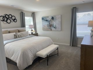A serene bedroom featuring a plush bed with neutral bedding, a modern abstract painting, decorative metal wall art, and soft grey curtains framing bright windows.