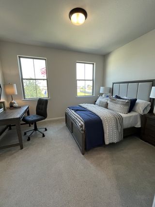 A cozy bedroom with a plush bed, elegant desk, and soft carpeting, bathed in natural light from two large windows.