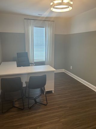 Model Home A modern home office with a sleek white desk, two black chairs, and warm wood flooring, complemented by chic lighting.