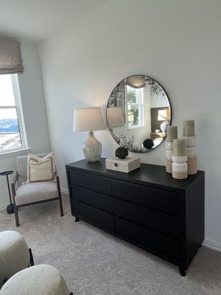 A cozy bedroom corner features a plush carpet, sleek black dresser, a round mirror, and elegant decor accents.
