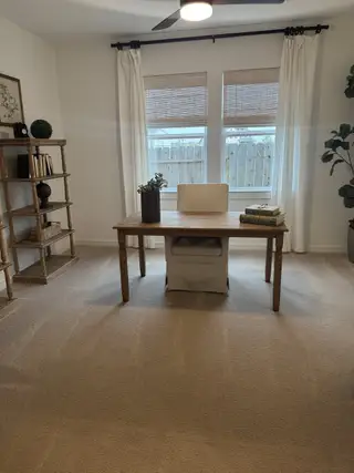 A cozy home office with a wooden desk, beige carpet, and ample natural light through large windows.