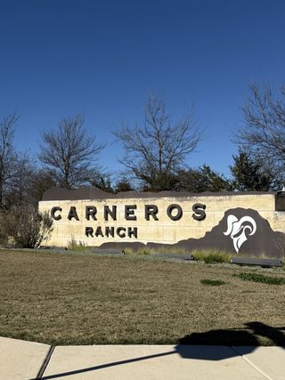 Street view Carneros Ranch by Coventry Homes in Leander, TX offers a welcoming entrance with its robust stone sign amidst natural landscaping.