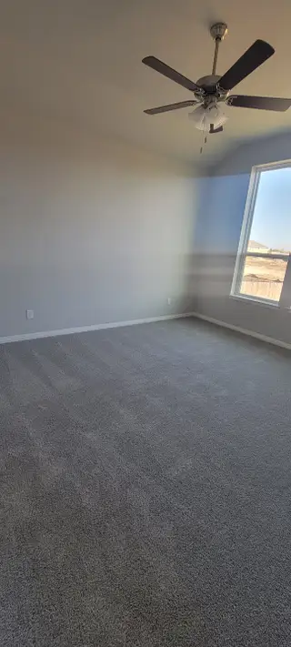 A spacious bedroom with plush carpeting, neutral walls, and a ceiling fan, featuring a large window for ample natural light.