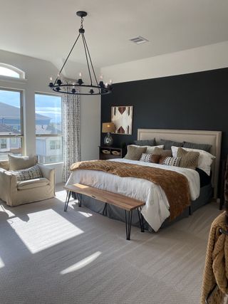 A cozy bedroom features a large bed, chic chandelier, and ample natural light with neutral tones and stylish decor.