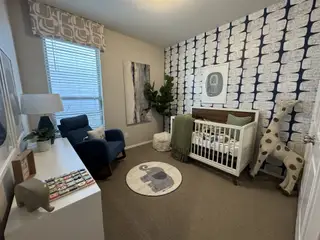 A cozy nursery featuring a crib, plush giraffe, patterned accent wall, and comfortable seating with natural light.