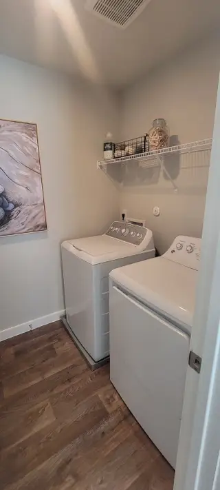 Model Home A cozy laundry room with wood flooring, featuring a washer, dryer, and overhead shelving for organized storage.