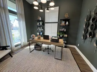 A modern home office with dark green walls, open shelving, and a sleek desk illuminated by a contemporary light fixture.
