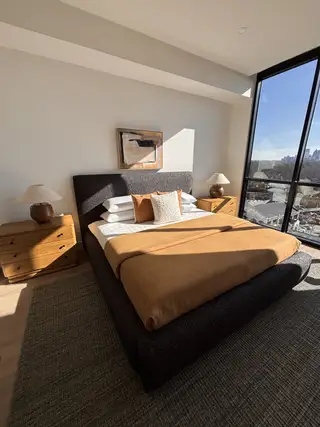 A cozy bedroom featuring a plush bed, warm-toned bedding, elegant nightstands, and a large window with city views.