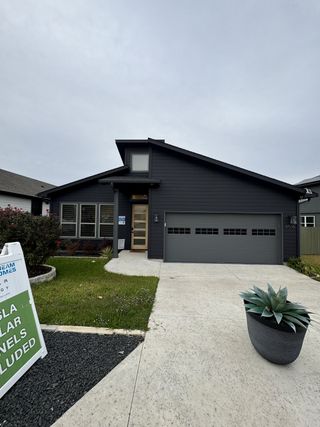 A sleek modern home with dark siding, large windows, and a landscaped front yard in Whisper Valley by AHA Dream Homes, LLC (Manor, TX).
