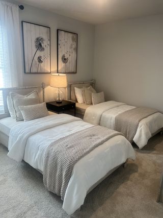 A charming guest bedroom features two identical twin beds with soft linens, a shared nightstand, and framed botanical prints above each bed.