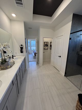 A modern bathroom with light tiled flooring, dual sinks, and a sleek glass shower, leading to a cozy bedroom.