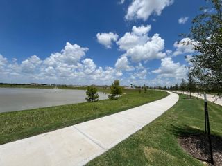 A scenic lakeside walking path in Miller's Pond by M/I Homes, Rosenberg, TX, with a vibrant blue sky and lush greenery.