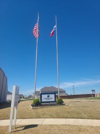 Entrance to Village on Main Street by Normandy Homes, marked by flags and landscaped sign, in Frisco, TX.