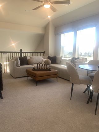 Model Home A cozy lounge area with plush seating, a stylish ottoman, and soft carpeting, illuminated by large windows and modern lighting.