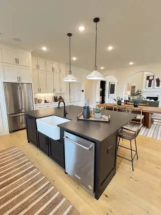 Model Home A modern kitchen with a dark island, farmhouse sink, and white cabinetry, featuring sleek lighting and stylish decor.