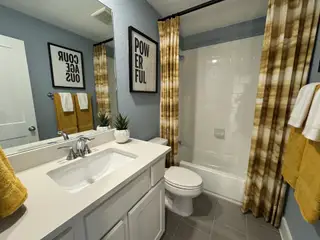 A modern bathroom with sleek white cabinetry, gray tiles, and stylish wall art, enhanced by warm-toned towels and curtains.