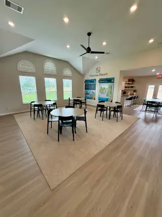 An inviting community room with round tables, wood flooring, large windows, and ceiling fan. Bright and welcoming ambiance.