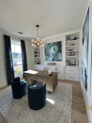 A cozy home office featuring elegant artwork, built-in shelving, plush carpeting, and modern chandelier lighting.