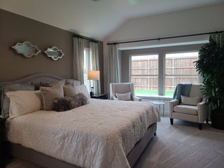 A cozy bedroom with elegant decor, featuring a plush bed, armchairs, and large windows overlooking a fenced backyard.