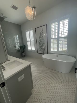 Model Home A chic bathroom featuring a freestanding tub, gray vanity, hexagonal tile flooring, and large windows with shutters.
