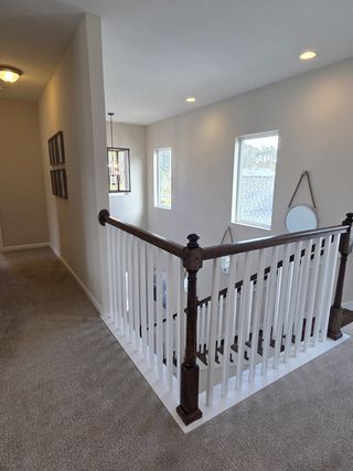A bright hallway with carpeted floors, elegant railing, and modern lighting fixtures, featuring large windows and wall decor.