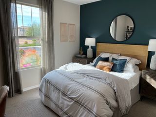 A cozy bedroom with a teal accent wall, round mirror, and plush bedding. Bright window view enhances the serene ambiance.