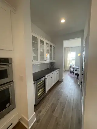 A sleek kitchen hallway with modern white cabinetry, dark countertops, and a wine cooler, leading to a bright dining area.