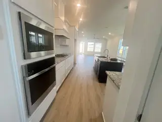 A sleek kitchen with stainless steel appliances, white cabinetry, a dark island with granite countertops, and light wood flooring extending into a bright living space.