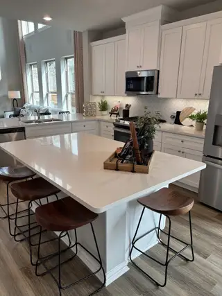 A modern kitchen with white cabinets, a large island, and barstools, featuring elegant lighting and wood flooring.