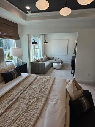 A cozy bedroom with a plush bed, textured blankets, modern lighting, and a view into a chic, light-filled sitting area.