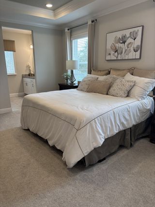 A cozy bedroom with soft carpeting, elegant tray ceiling, and neutral decor, leading to a bright vanity area.