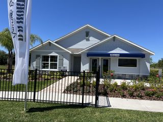 A charming single-story model home with a blue awning, white siding, and lush landscaping in Berry Bay by D.R. Horton (Wimauma, FL).
