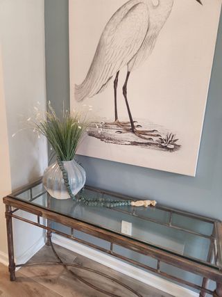 A chic entryway with a glass-top table, decorative vase, and elegant bird artwork on a soft gray wall.