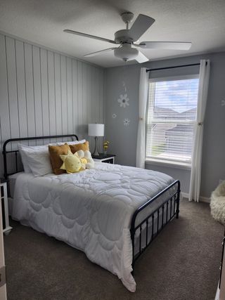 Cozy bedroom with a quaint metal bed, floral bedding, and cheerful sun pillow. Light gray panel walls and soft carpeting.
