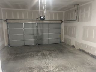 A bare garage with a gray metal garage door, exposed drywall, a concrete floor, and an overhead door mechanism.
