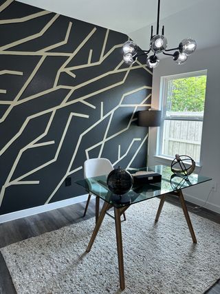 Model Home A modern office with a geometric accent wall, glass desk, contemporary lighting, and plush rug.