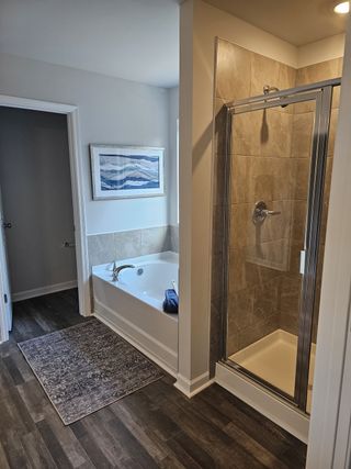 A bathroom features a soaking tub with tile surround, a glass-enclosed shower, dark wood-look flooring, and a decorative rug, creating a blend of comfort and style.
