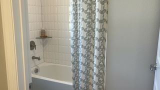 Model Home A cozy bathroom with a white tiled bathtub, elegant shower curtain, and glass shelf for toiletries.