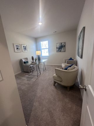 A cozy home office with beige carpeting, a glass desk, plush seating, and modern artwork on the walls.