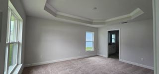 A bright room with a tray ceiling, plush carpeting, and ample natural light from the windows, leading to an adjoining bathroom.