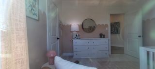 A cozy bedroom with soft pink scalloped walls, a white dresser, round mirror, and playful decor accents.
