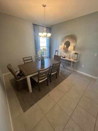 Model Home An elegant dining room with a wooden table, wicker chairs, and chic chandelier, complemented by soft gray walls and tiled floor.