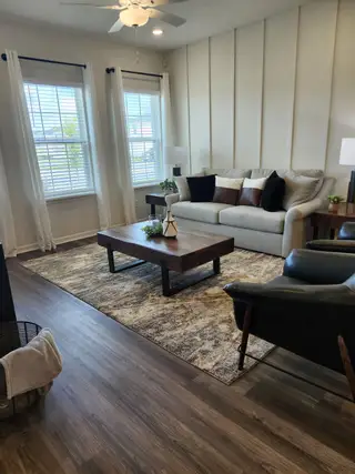 A cozy living room with a plush sofa, wooden coffee table, and large windows with sheer drapes, on a textured rug.